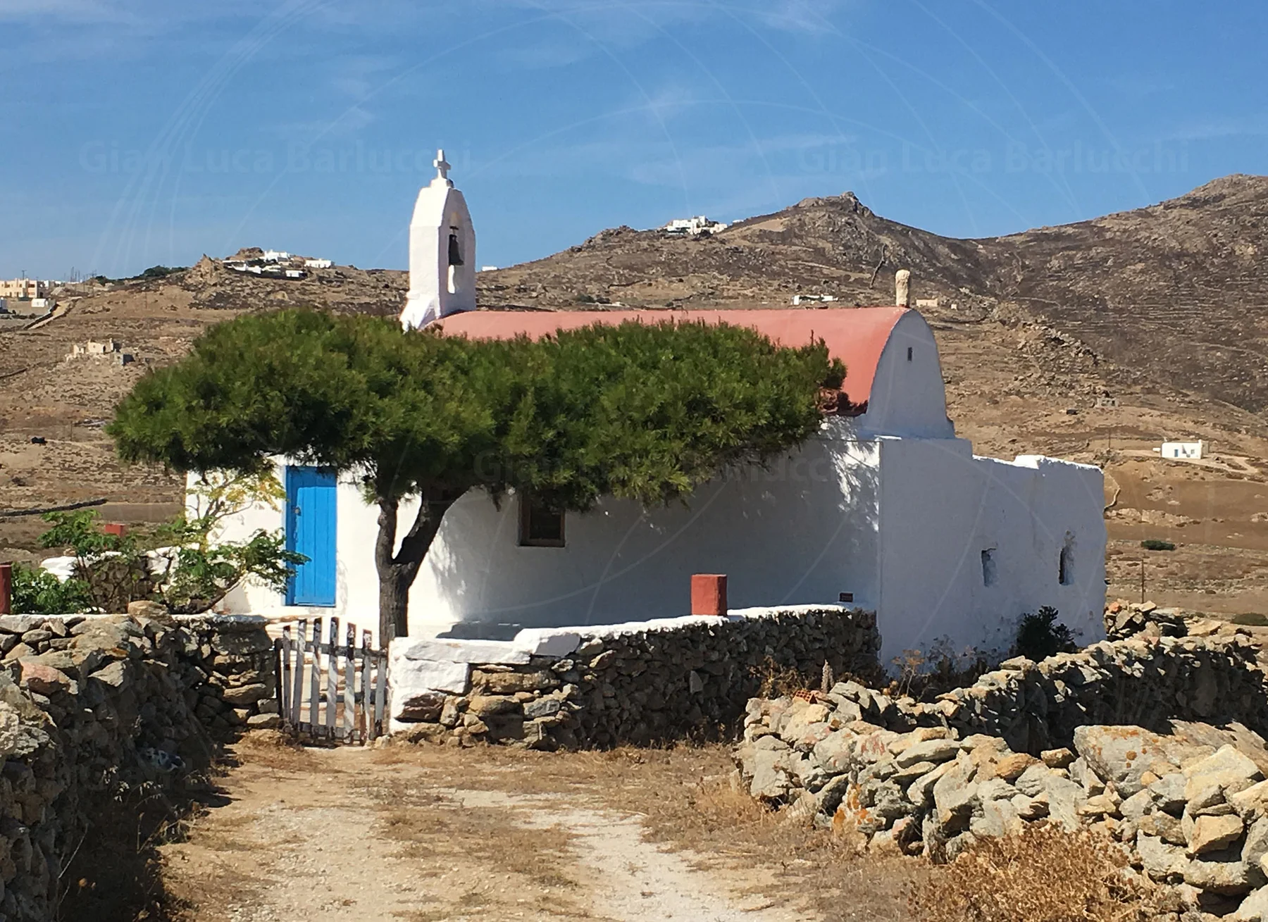 Small white church with a red roof and blue door among stone walls on Mykonos