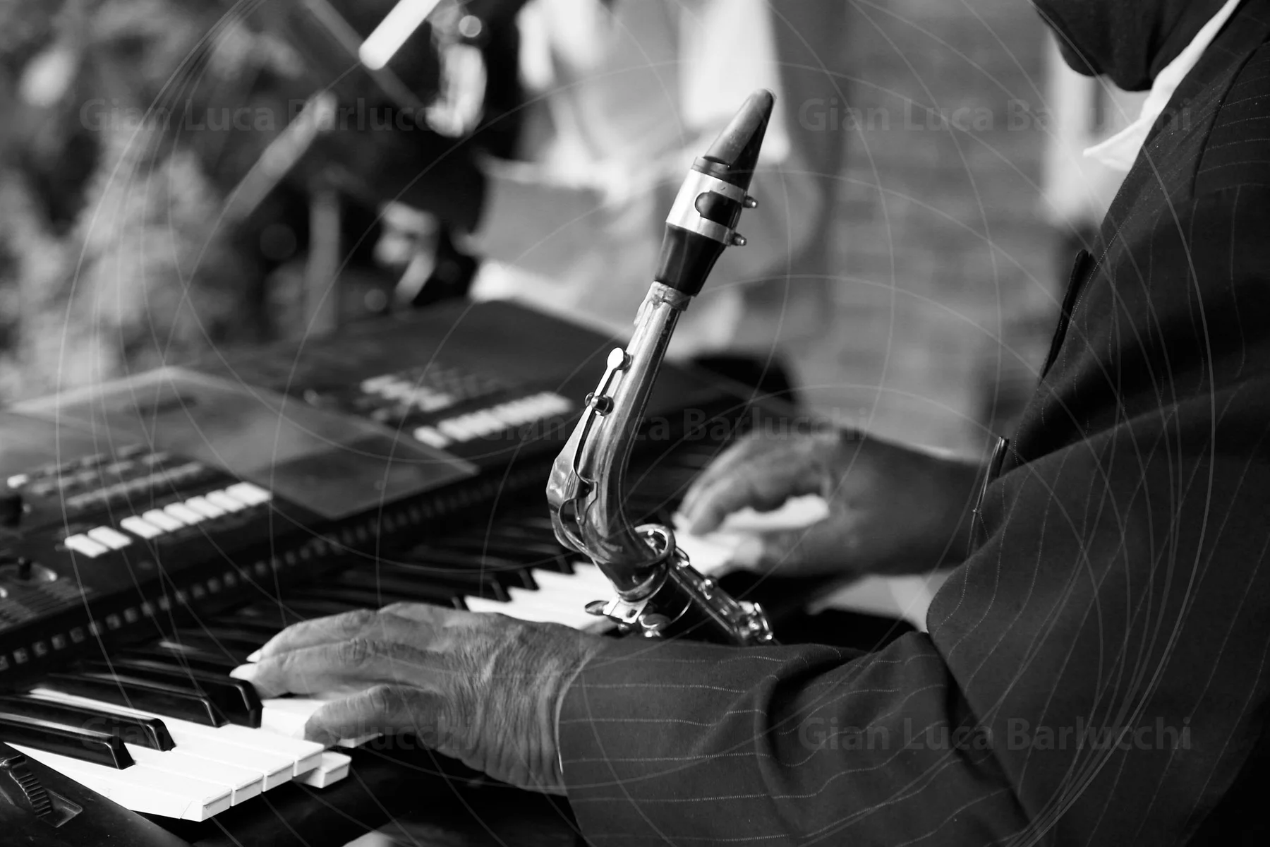 Black-and-white detail of a musician playing a keyboard while holding a saxophone