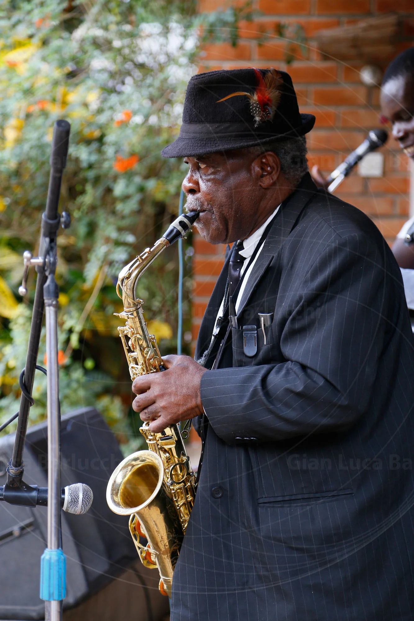 Musician in a dark suit and hat playing saxophone beside a microphone
