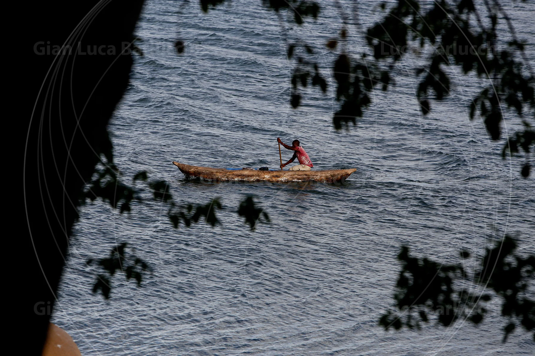 Person paddling a wooden canoe on rippled water, framed by branches